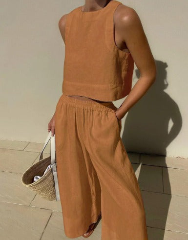 Frau in beigefarbenem Leinen-Zweiteiler mit ärmellosem Top und weiter Hose, hält eine Strohtasche. Sommermode, minimalistischer Stil.