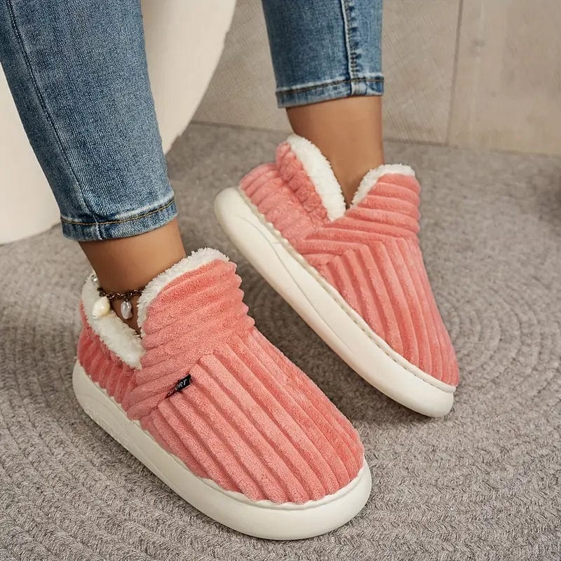 Damen tragen rosa, plüschige Hausschuhe mit weißem Futter, kombiniert mit blauen Jeans. Bequeme, warme Slipper auf grauem Teppichboden.