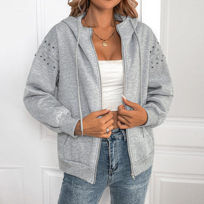 Frau in grauem Kapuzenpullover mit Nieten, weißem Top und blauer Jeans vor weißer Tür. Modetrend, lässiger Stil, Damenbekleidung.