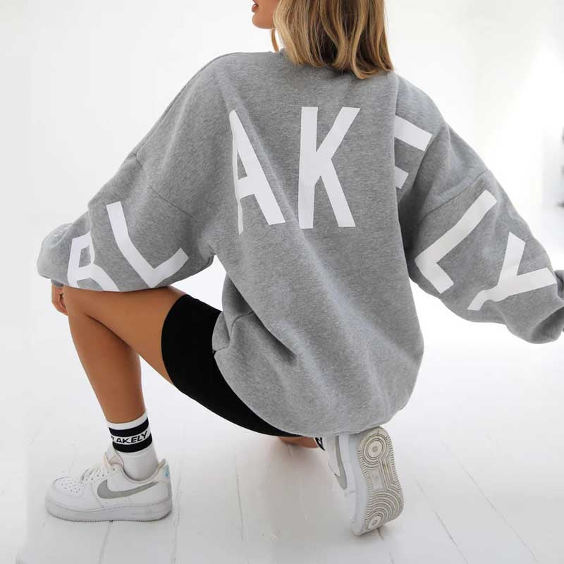 Frau in grauem Blakely-Sweatshirt und schwarzen Shorts, kniend auf weißem Boden, trägt weiße Sneakers. Sportliche Mode, lässiger Stil.