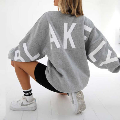 Frau in grauem Blakely-Sweatshirt und schwarzen Shorts, kniend auf weißem Boden, trägt weiße Sneakers. Sportliche Mode, lässiger Stil.