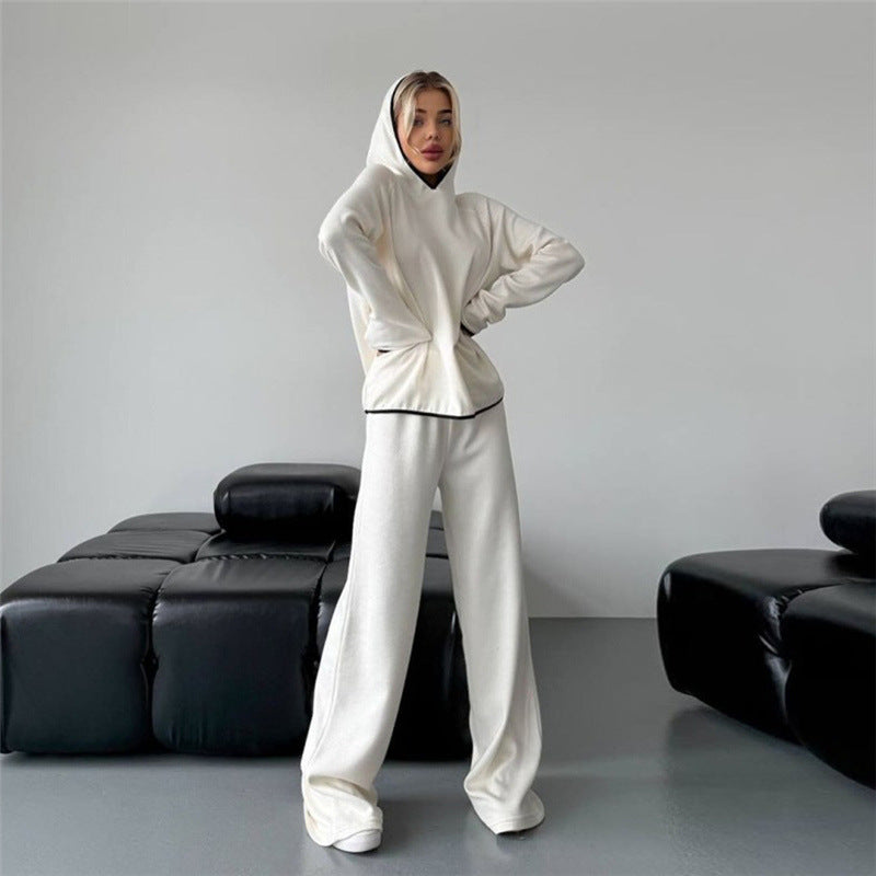 Frau in weißem Hoodie und Jogginghose posiert in modernem Wohnzimmer mit schwarzen Sofas. Bequeme Freizeitmode, minimalistisches Interieur.