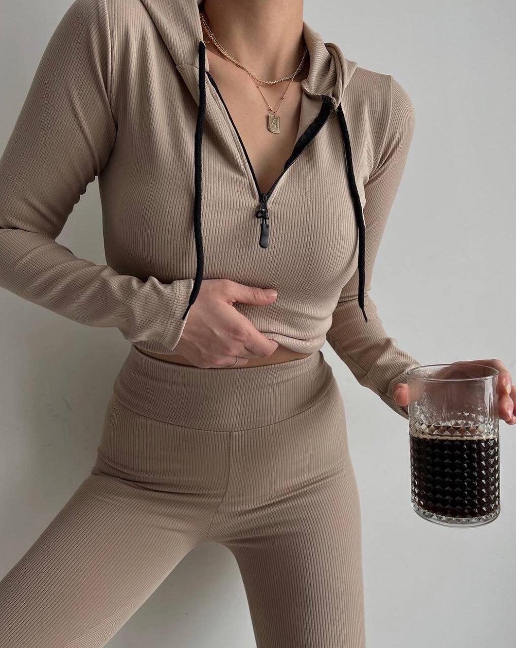 Frau in beige geripptem Trainingsanzug hält Glas mit dunklem Getränk. Sportmode, athleisure, Fitness-Outfit, modischer Hoodie, gesunder Lebensstil.