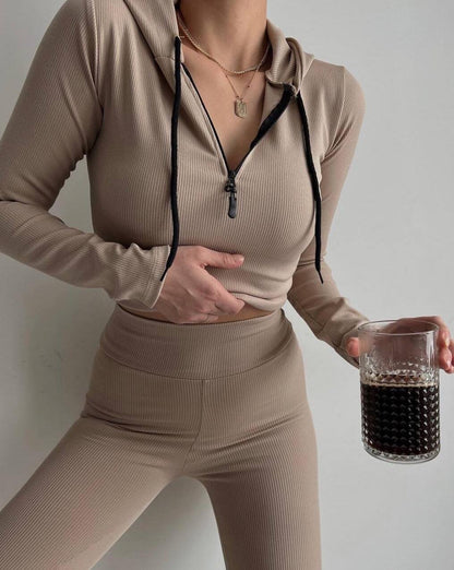 Frau in beige geripptem Trainingsanzug hält Glas mit dunklem Getränk. Sportmode, athleisure, Fitness-Outfit, modischer Hoodie, gesunder Lebensstil.