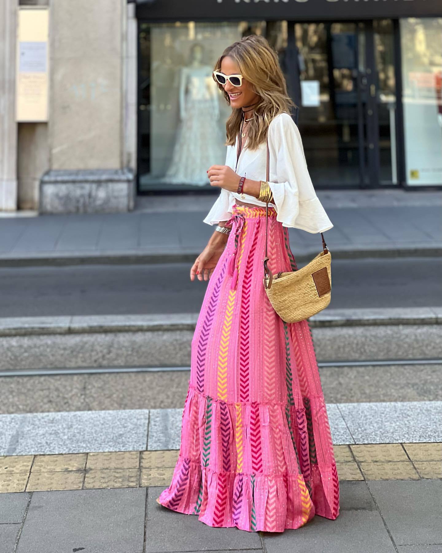 Frau in modischem Sommeroutfit mit weißer Bluse und pinkem Maxirock, Sonnenbrille und Strohtasche, spaziert auf städtischem Gehweg.