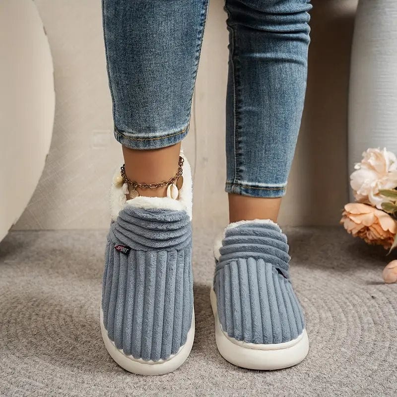 Graue, gestreifte Hausschuhe aus Plüsch an Füßen in blauen Jeans, auf Teppichboden. Bequeme, warme Damen-Hausschuhe für den Winter.