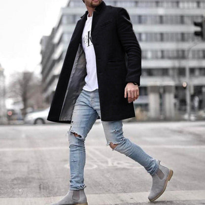 Mann in schwarzem Mantel, zerrissenen Jeans und grauen Chelsea-Stiefeln überquert Straße in urbaner Umgebung. Modetrend, Herrenmode, Streetstyle.