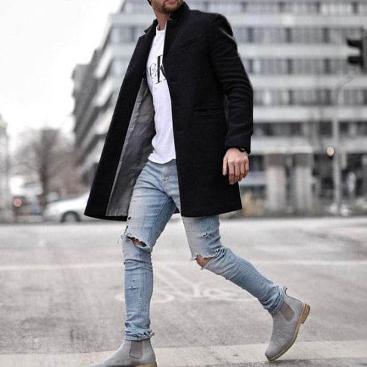 Mann in schwarzem Mantel, zerrissenen Jeans und grauen Chelsea-Stiefeln überquert Straße in urbaner Umgebung. Modetrend, Herrenmode, Streetstyle.