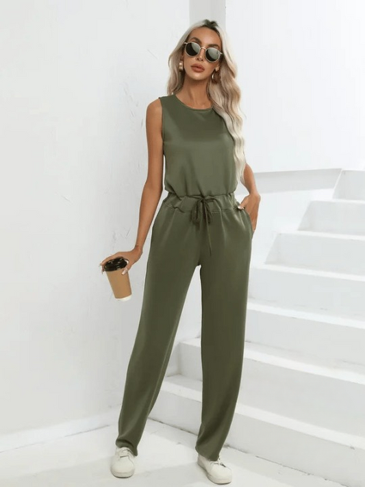 Frau in olivgrünem Jumpsuit mit Sonnenbrille und Kaffeebecher, steht vor weißen Treppen. Modetrend, lässiger Stil, Sommeroutfit.