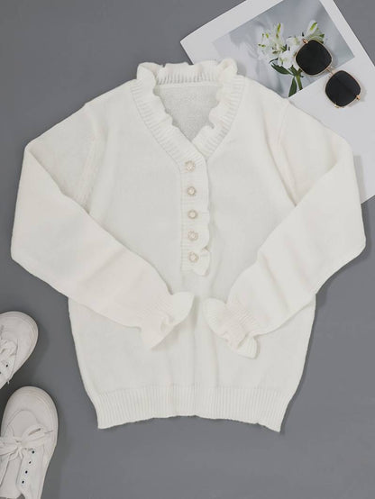 Weiße Strickjacke mit Rüschen und Perlenknöpfen auf grauem Hintergrund, neben weißen Sneakers und Sonnenbrille mit Blumenfoto.