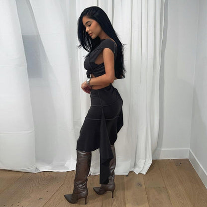 Frau in elegantem schwarzen Kleid und braunen Stiefeln posiert vor weißen Vorhängen auf Holzboden. Mode, Stil, Abendkleid, Damenmode.