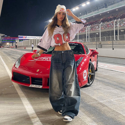 Frau in weiter Jeans und Crop-Top posiert vor rotem Ferrari auf Rennstrecke bei Nacht. Motorsport, Mode, Luxusauto, Nachtaufnahme.