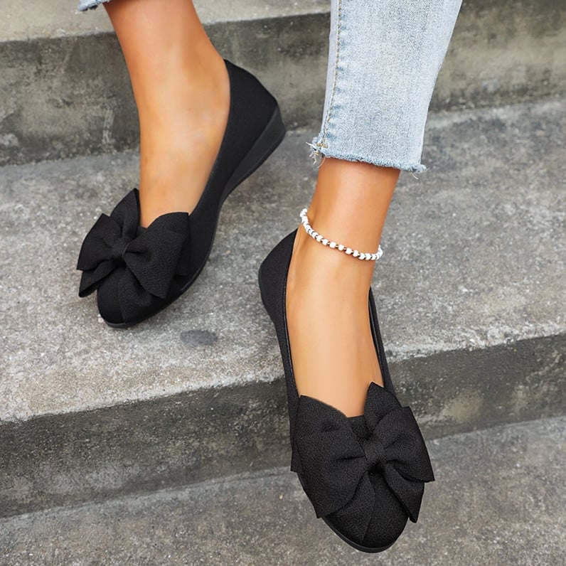 Damenfüße in schwarzen Ballerinas mit großer Schleife, kombiniert mit hellblauen Jeans und silbernem Fußkettchen, auf grauer Treppe.