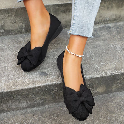 Damenfüße in schwarzen Ballerinas mit großer Schleife, kombiniert mit hellblauen Jeans und silbernem Fußkettchen, auf grauer Treppe.