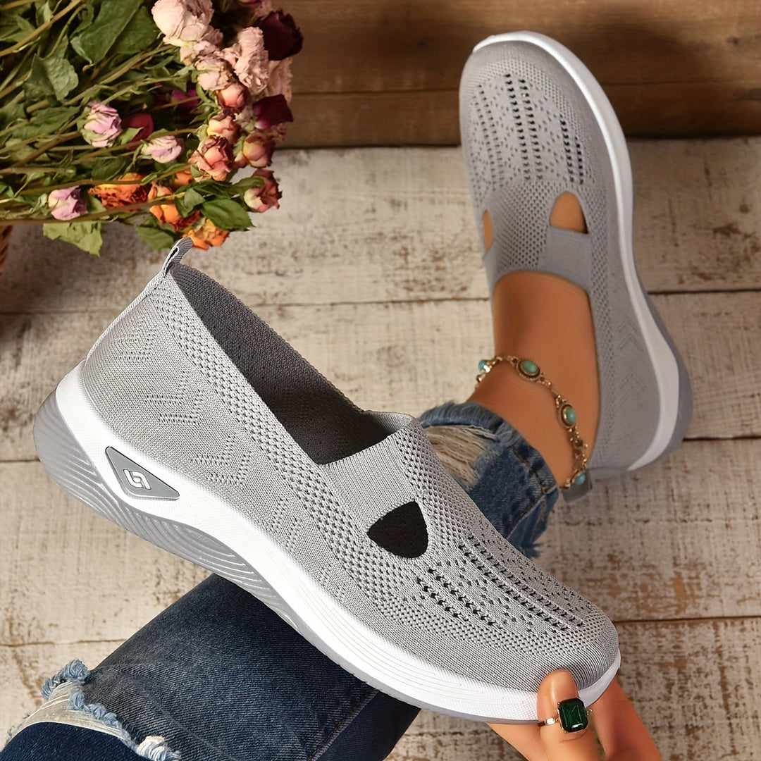 Graue Damen-Sneaker aus atmungsaktivem Strickmaterial, getragen von Person mit Jeans, auf Holzboden, neben einem Blumenstrauß.