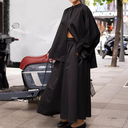 Frau in schwarzem, weitem Outfit steht neben einem Roller auf einer städtischen Straße. Modetrend, urbaner Stil, minimalistisches Design.