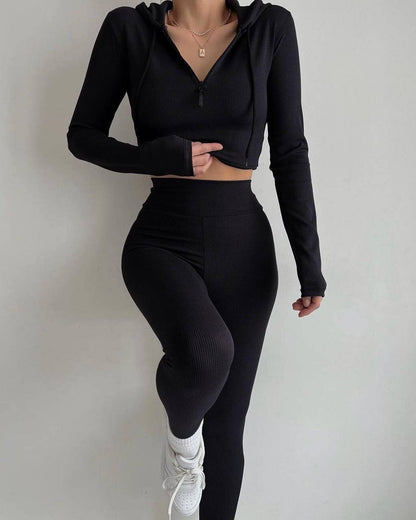 Frau in schwarzem Sport-Set mit Kapuzenjacke und Leggings, posiert vor weißem Hintergrund. Fitnessmode, Athleisure, Sportbekleidung.