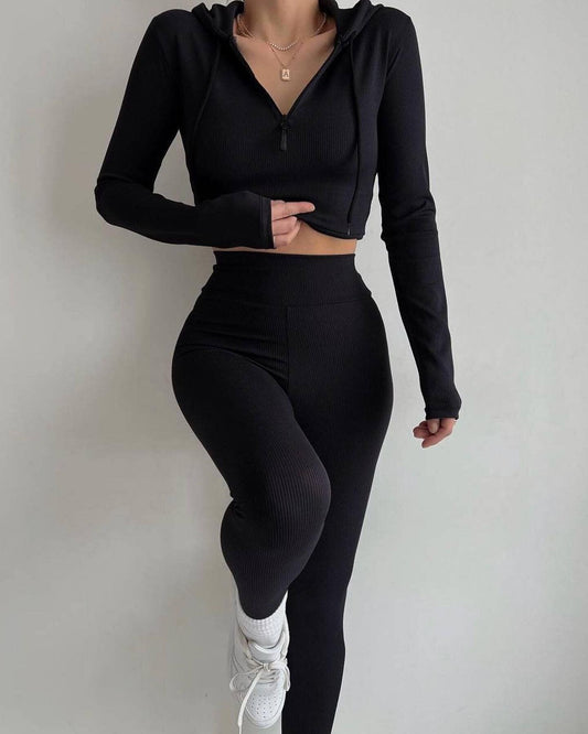 Frau in schwarzem Sport-Set mit Kapuzenjacke und Leggings, posiert vor weißem Hintergrund. Fitnessmode, Athleisure, Sportbekleidung.
