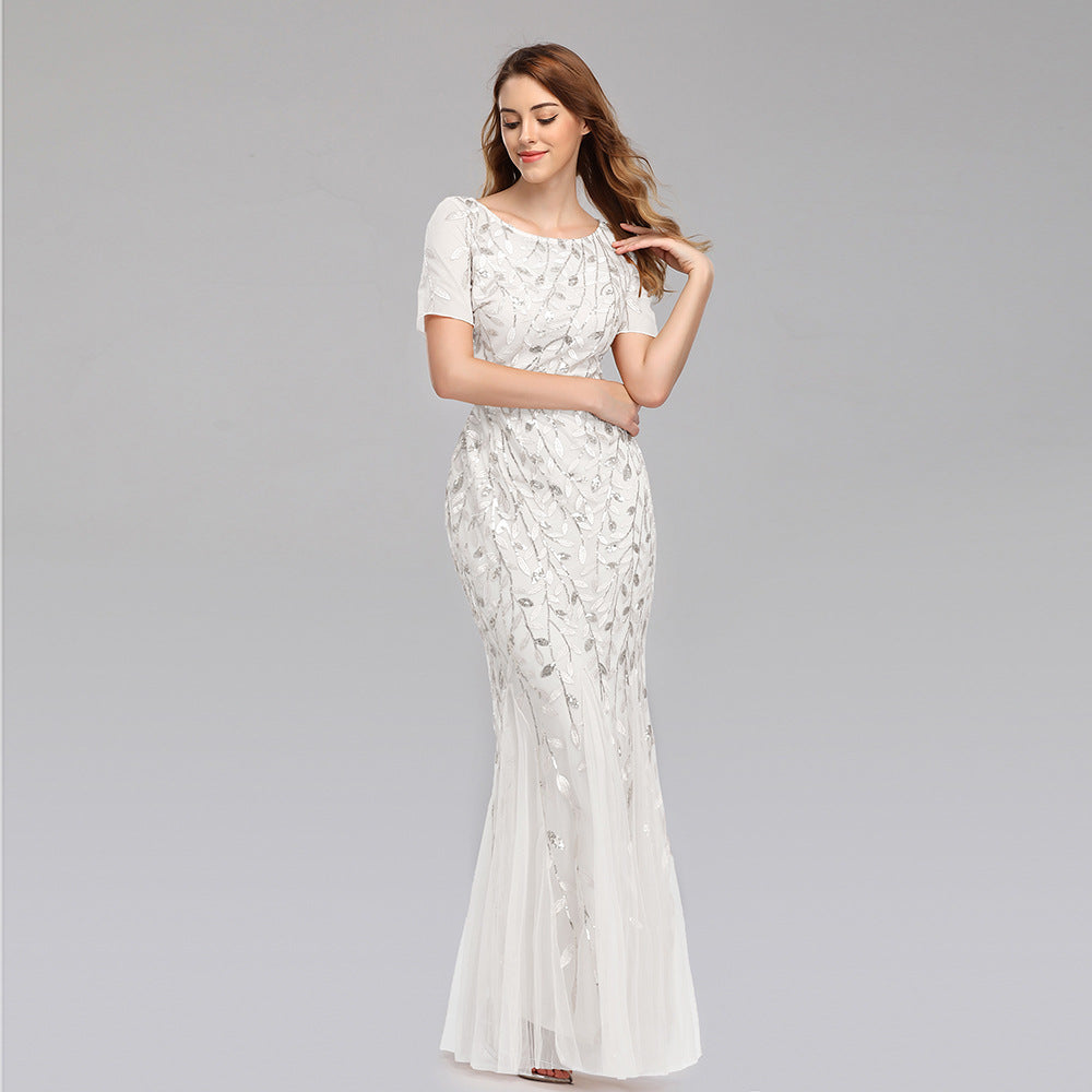 Frau in elegantem, weißem Abendkleid mit floralen Stickereien, lächelt und posiert vor grauem Hintergrund. Perfekt für Hochzeiten oder formelle Anlässe.