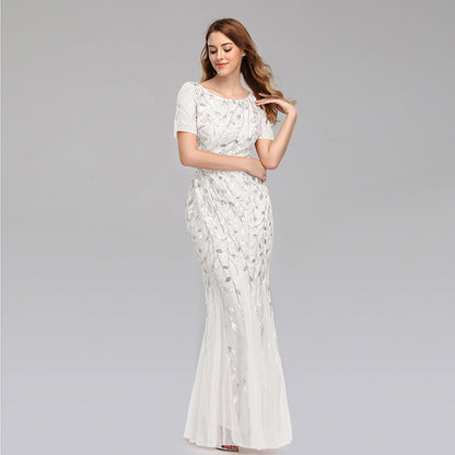 Frau in elegantem, weißem Abendkleid mit floralen Stickereien, lächelt und posiert vor grauem Hintergrund. Perfekt für Hochzeiten oder formelle Anlässe.