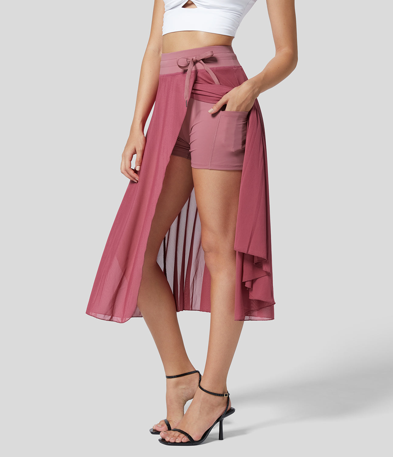 Frau in rosa Maxirock mit Shorts, weißem Crop-Top und schwarzen High Heels. Modetrend, Sommeroutfit, stilvolle Damenbekleidung.