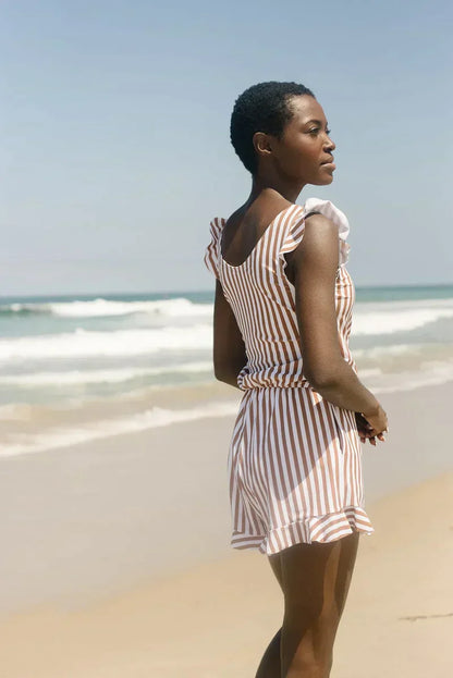 Frau am Strand in gestreiftem Sommerkleid, blickt aufs Meer. Sonniger Tag, Wellen im Hintergrund. Perfekt für Strandmode und Sommerurlaub.