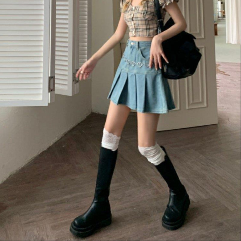 Junge Frau in kariertem Top und Jeansrock mit Herzgürtel, trägt schwarze Stiefel und weiße Kniestrümpfe, posiert in modernem Innenraum.