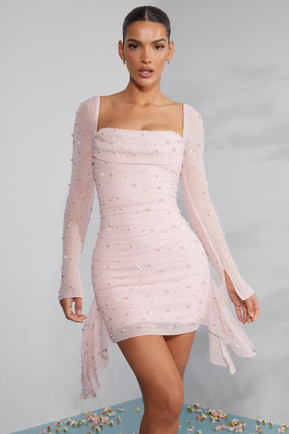 Frau in elegantem, rosa Paillettenkleid mit langen Ärmeln und asymmetrischem Saum, posiert vor neutralem Hintergrund. Abendmode, Damenkleidung.