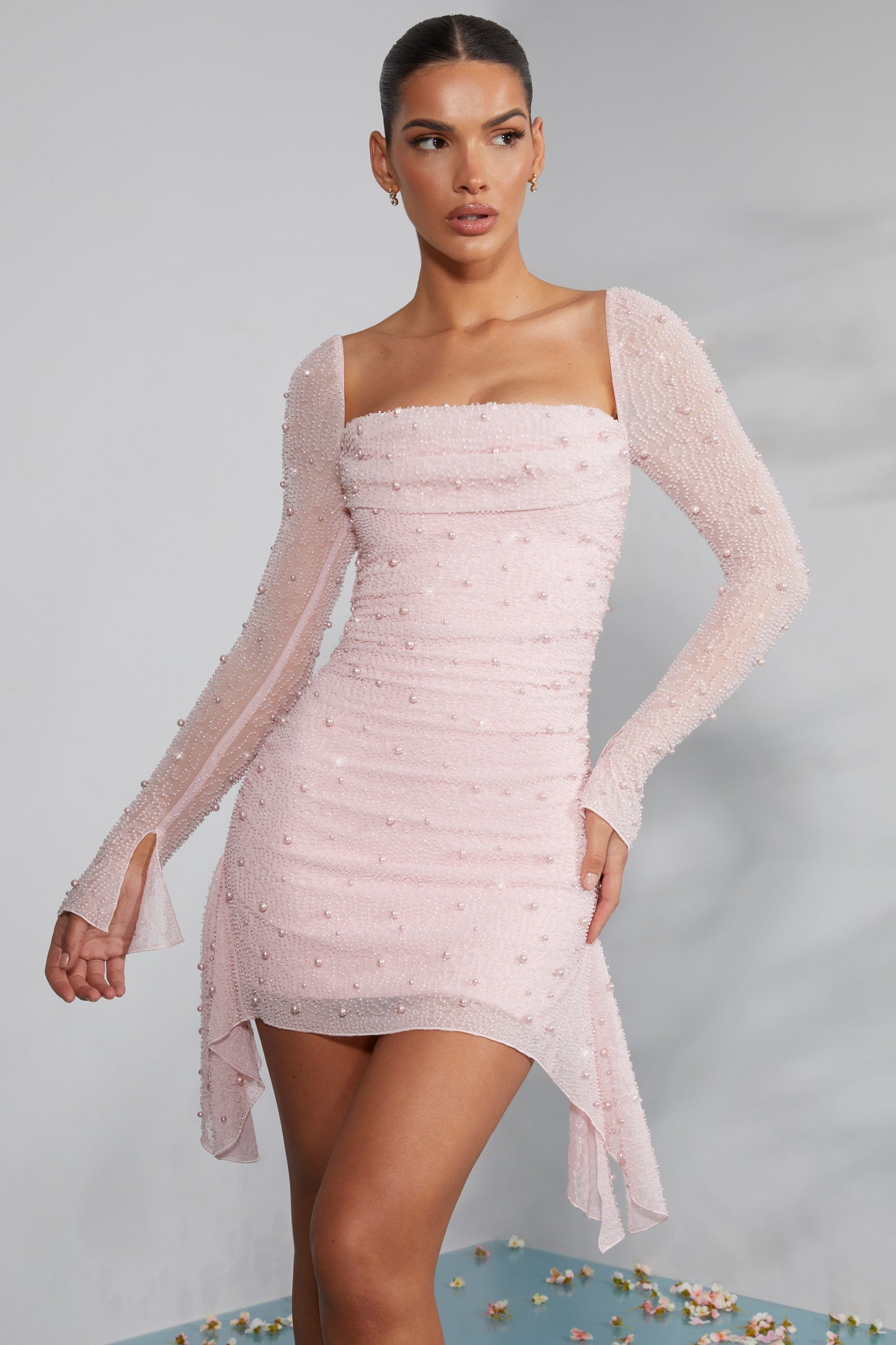 Frau in elegantem, rosa Minikleid mit langen Ärmeln und Perlendetails, posiert vor grauem Hintergrund. Modetrend, Abendmode, stilvolles Design.