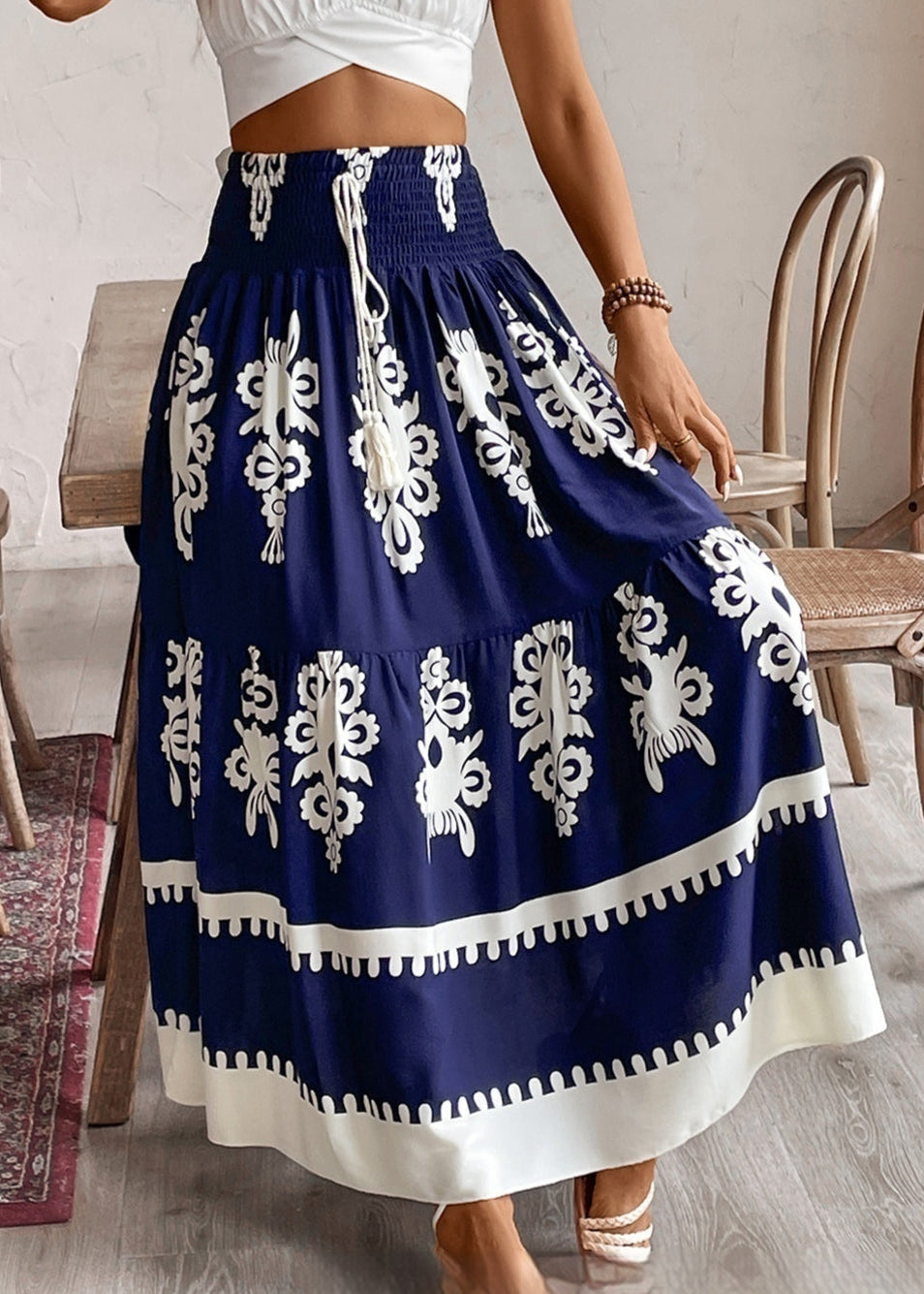 Frau in weißem Top und blau-weißem Maxirock mit floralem Muster, steht in einem Raum mit Holzboden und Stühlen. Boho-Chic, Sommermode, Damenbekleidung.