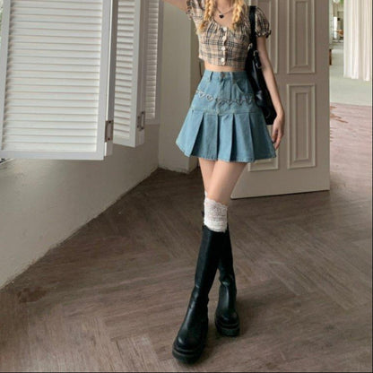 Junge Frau in kariertem Crop-Top und Jeansrock mit Herzketten-Detail, trägt hohe schwarze Stiefel und weiße Socken, steht in modernem Raum.