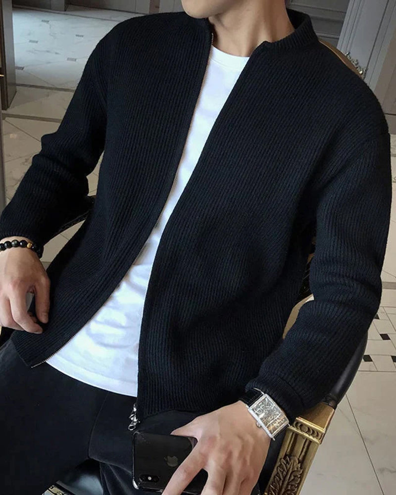 Mann in schwarzer Strickjacke und weißem T-Shirt, hält Smartphone, trägt Armbanduhr. Moderne Herrenmode, lässiger Stil, Innenaufnahme.
