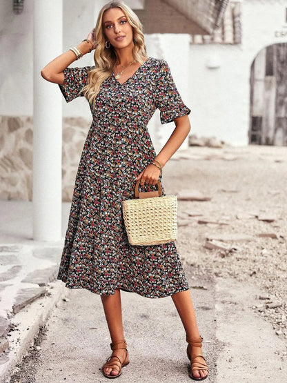Frau in geblümtem Sommerkleid mit Strohtasche, steht auf gepflastertem Weg vor weißem Gebäude. Mode, Sommerkleid, Strohtasche, Damenmode.
