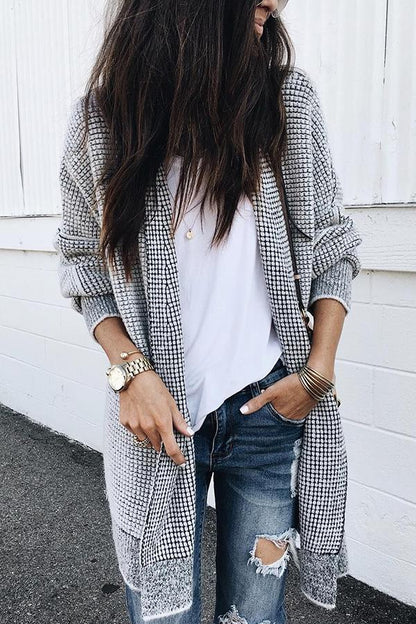 Frau in grauem Strickcardigan, weißem T-Shirt und zerrissenen Jeans vor weißer Wand. Modischer Streetstyle-Look, lässig und trendig.
