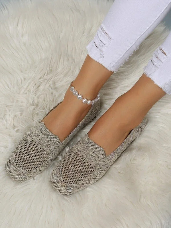 Damenfüße in beigen Strick-Loafers auf weißem Fellteppich, weiße zerrissene Jeans, Perlenknöchelband. Modische Damen-Schuhe, gemütlicher Stil.