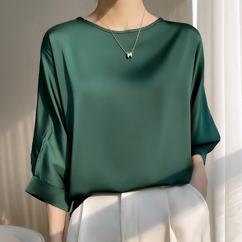 Dame in eleganter, grüner Seidenbluse mit Dreiviertelärmeln und weißem Rock, trägt eine Halskette. Perfekt für stilvolle Büro- oder Abendmode.