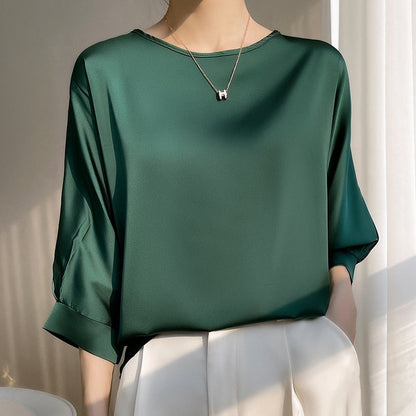 Dame in eleganter, grüner Seidenbluse mit Dreiviertelärmeln und weißem Rock, trägt eine Halskette. Perfekt für stilvolle Büro- oder Abendmode.