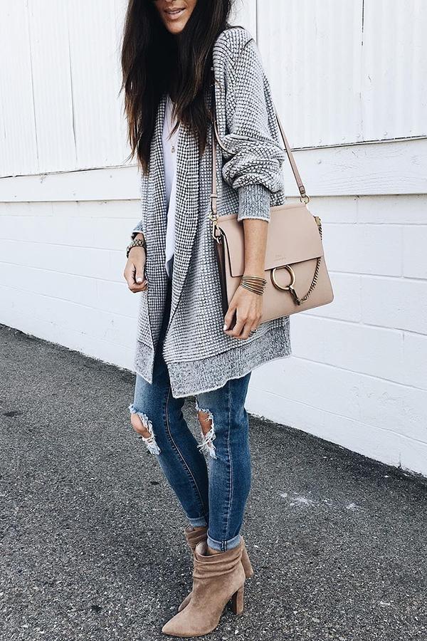 Frau in modischem Herbst-Outfit mit grauem Cardigan, zerrissenen Jeans, beigen Stiefeletten und beiger Umhängetasche vor weißer Wand.