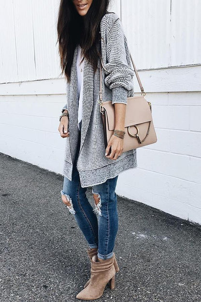 Frau in modischem Herbst-Outfit mit grauem Cardigan, zerrissenen Jeans, beigen Stiefeletten und beiger Umhängetasche vor weißer Wand.