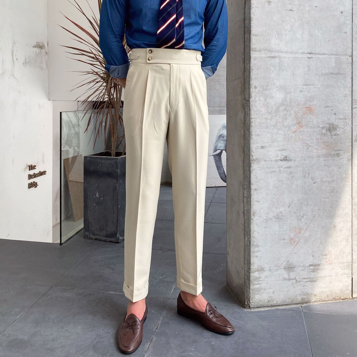Mann in beiger Bundfaltenhose, blauem Hemd und gestreifter Krawatte. Elegante braune Loafer, moderne Innenarchitektur im Hintergrund.