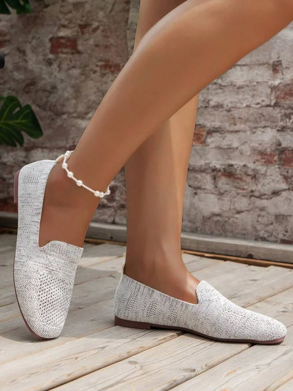 Weiße, gestrickte Damen-Loafer auf Holzboden, kombiniert mit einer Perlenfußkette. Perfekte Sommerschuhe für stilvolle Freizeitoutfits.