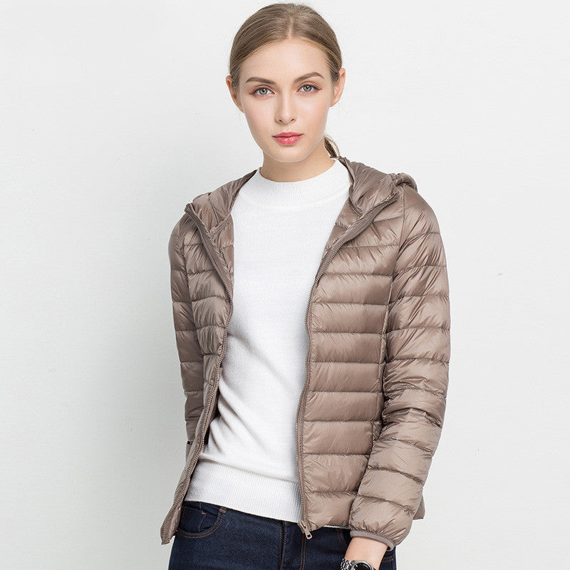Frau in beiger Daunenjacke mit Kapuze, weißem Pullover und blauer Jeans vor weißem Hintergrund. Modetrend, Winterjacke, Damenbekleidung.