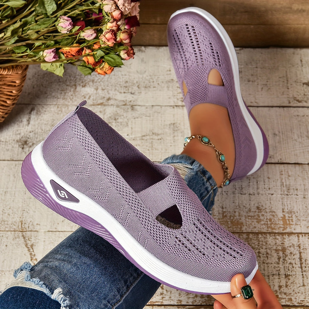 Lila Damen-Sneaker aus Strickmaterial, atmungsaktiv, mit weißer Sohle, getragen von Person in Jeans, auf Holzboden mit Blumen im Hintergrund.