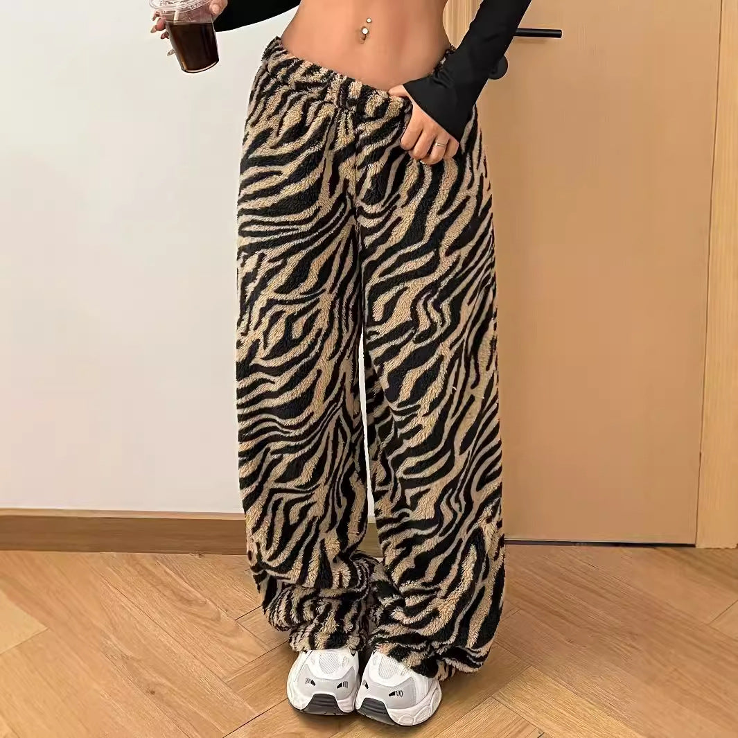 Frau in Zebra-Muster Plüschhose, schwarze Langarm-Crop-Top, weiße Sneaker, hält Getränk. Modetrend, bequeme Kleidung, Streetstyle.