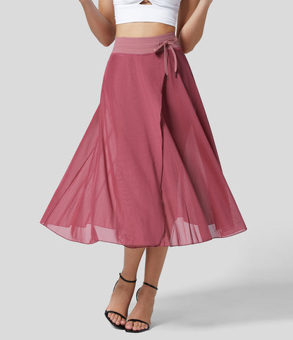 Frau in rosa Chiffon-Midirock mit Schleife, weißem Crop-Top und schwarzen Sandaletten. Elegante Damenmode, Sommeroutfit, stilvoller Look.