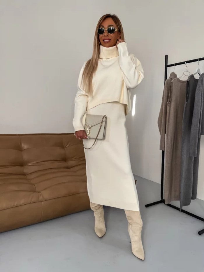 Frau in cremefarbenem Rollkragenpullover und Rock, trägt Sonnenbrille und Stiefel, steht neben braunem Sofa und Kleiderständer mit Kleidung.