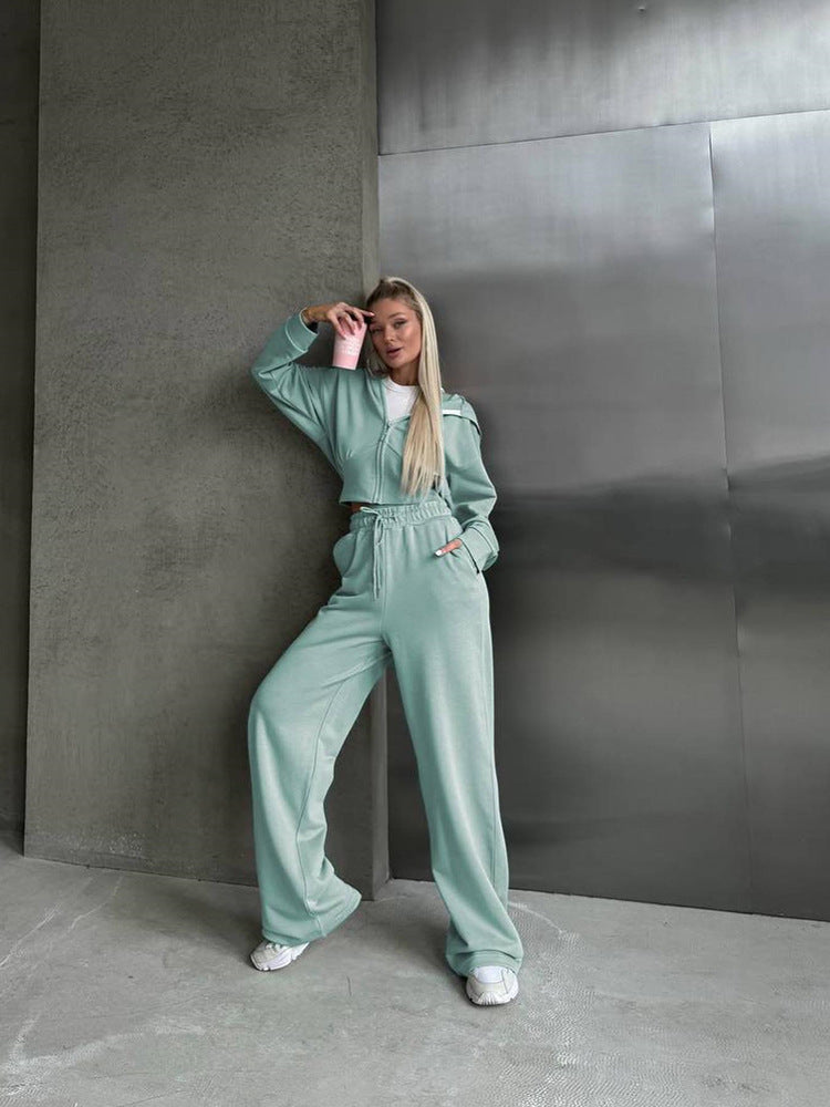 Frau in mintgrünem Jogginganzug lehnt an Betonwand, hält rosa Becher. Modetrend, Freizeitkleidung, urbaner Stil, bequeme Mode.