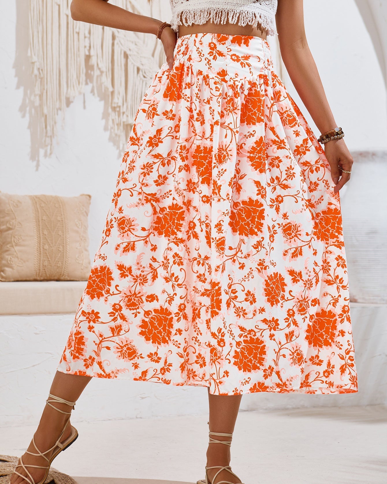 Frau in weißem, ärmellosem Oberteil und langem, orange-weiß geblümtem Rock, stehend in modernem, boho-inspiriertem Raum mit Makramee-Dekor.