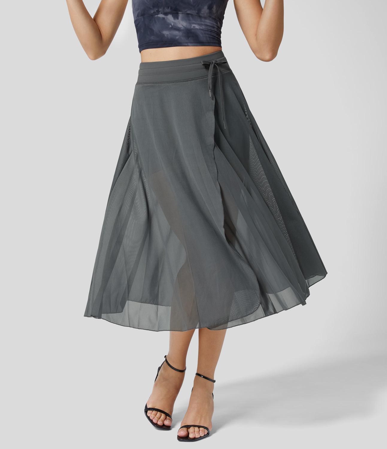 Damen in grauem, transparentem Chiffon-Wickelrock, knielang, kombiniert mit schwarzem Top und Sandalen. Elegante Sommermode, stilvoll und luftig.