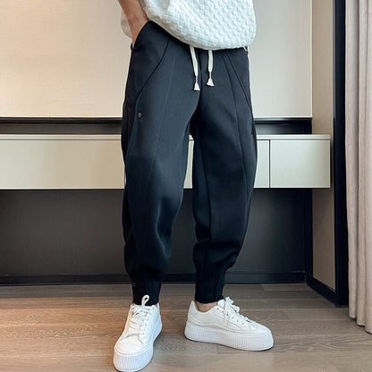 Person trägt schwarze Jogginghose und weiße Sneaker, steht auf Holzboden vor weißer Wand. Moderne Freizeitmode, sportlicher Look, Herrenbekleidung.
